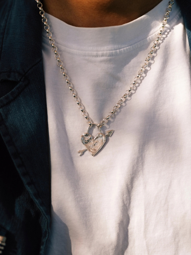 Medium heart arrow necklace. A silver heart with an arrow through it on a chain worn by someone in a white t-shirt