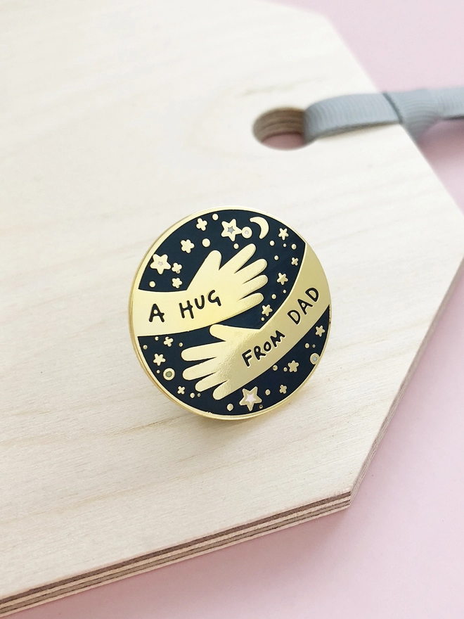 A navy blue and gold pin badge with a hugging arms design and the words “A hug from Dad” is placed on a wooden tag.