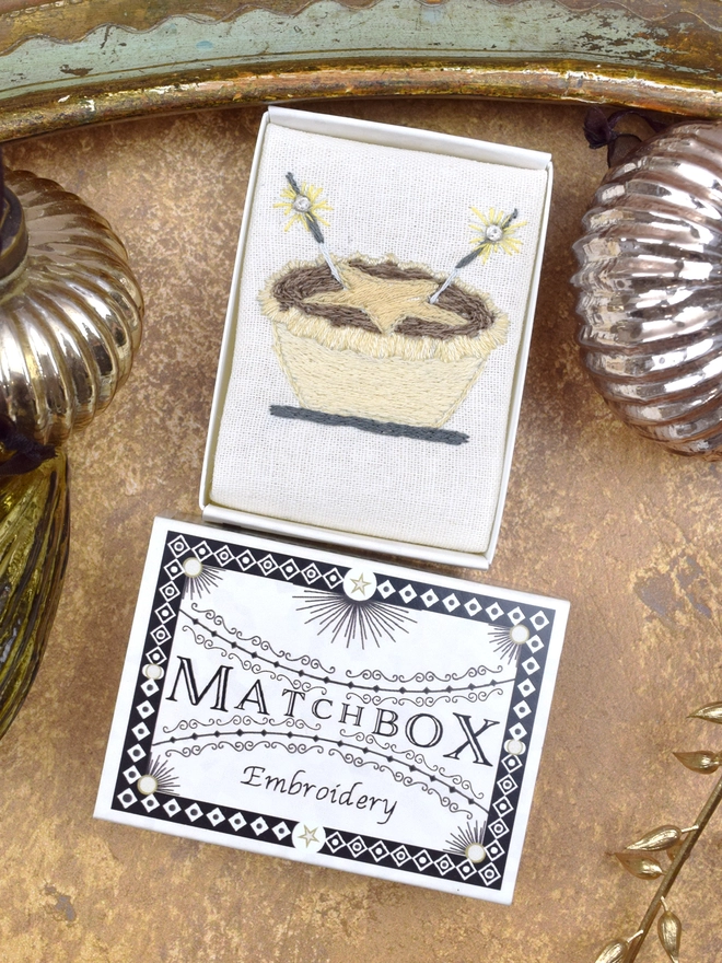 Mince Pie embroidery, inside a Matchbox displayed on a gold background with glass baubles.
