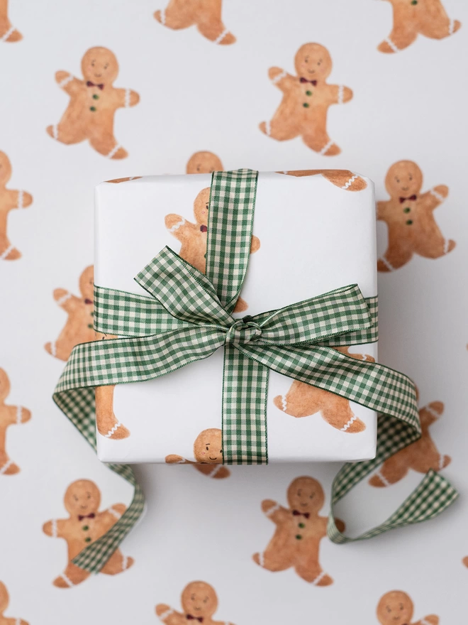 Gingerbread Man Gift Wrap featuring a repeat print of a hand painted Gingerbread Man illustration on the white base.  The Gift is tied with a Green Gingham Bow 