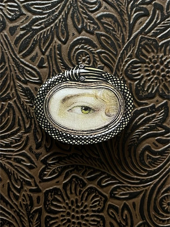 lovers eye brooch, the front is an illustration of a lovers eye with a snake like metal border, printed onto a wood brooch base, with a brass clasp at the back