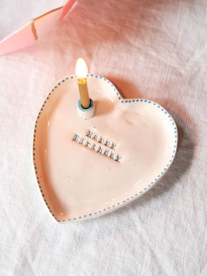 Handmade ceramic heart dish stamped with "Happy Birthday" featuring a blue dotted rim on a plain white table cloth with a birthday candle 