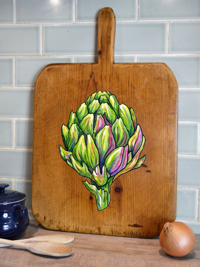 Wooden chopping board with handpainted design of an artichoke standing against a kitchen wall
