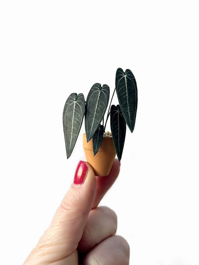 A miniature replica Warocqueanum Queen Anthurium paper plant ornament in a terracotta pot being held between 2 fingers against a white background