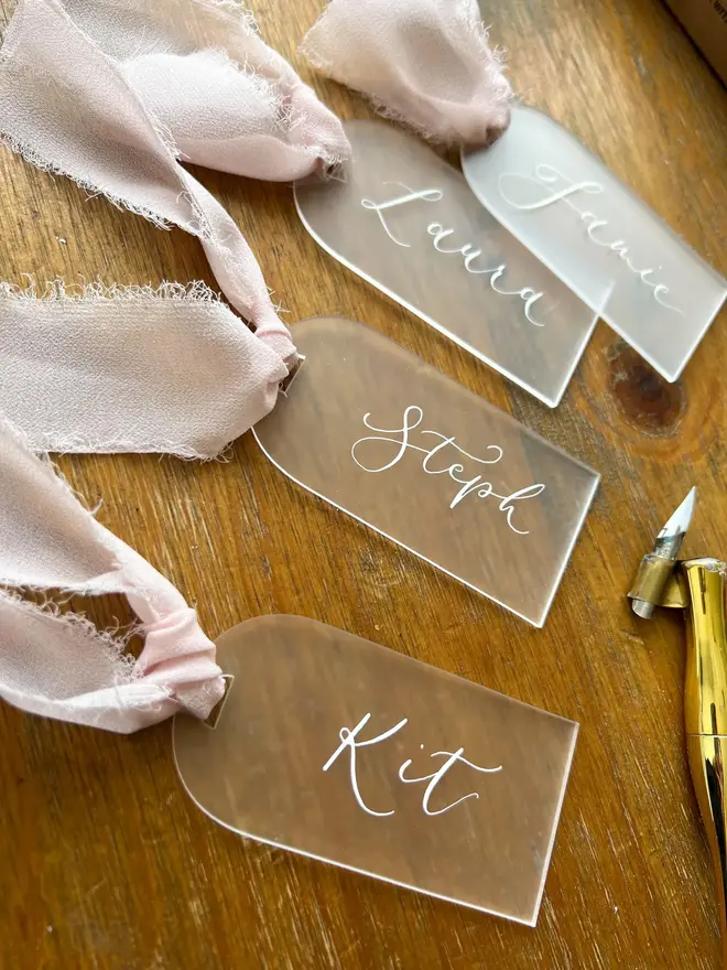 Personalised Calligraphy Frosted Acrylic Place Cards, frosted acrylic place cards with pink ribbons grouped together on a wooden table. 