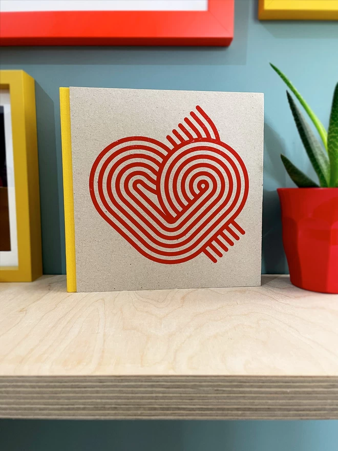 Stripy Heart design is screenprinted in magenta on a grey pasteboard sketchbook with a yellow fabric spine, stood on a plywood shelf with hints of framed pictures around. the wall is duck egg blue, a plant glimpsed to the side.