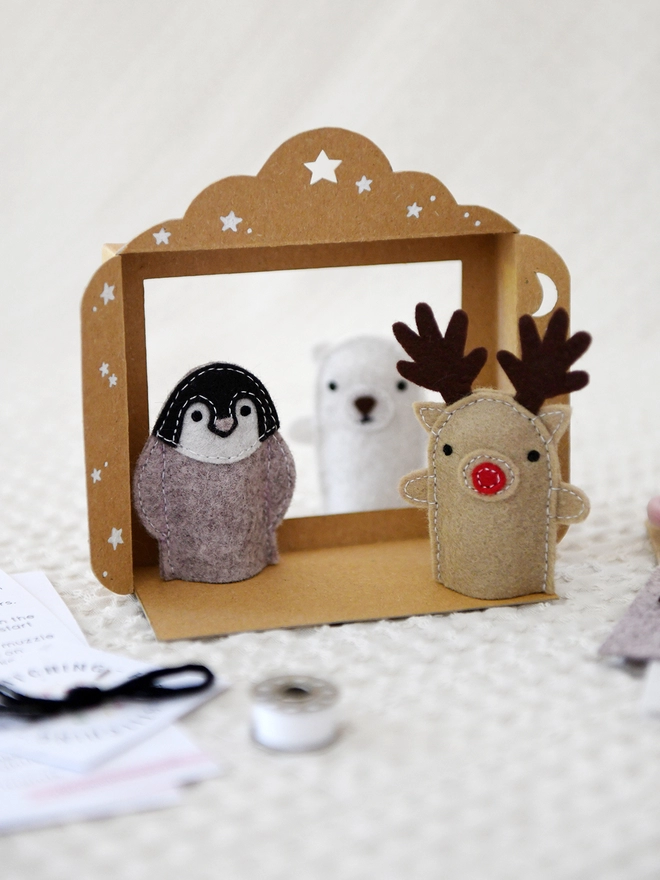 Three felt finger puppets, a reindeer, a penguin, and a polar bear, stand inside a small cardboard puppet theatre on an ivory fabric surface.