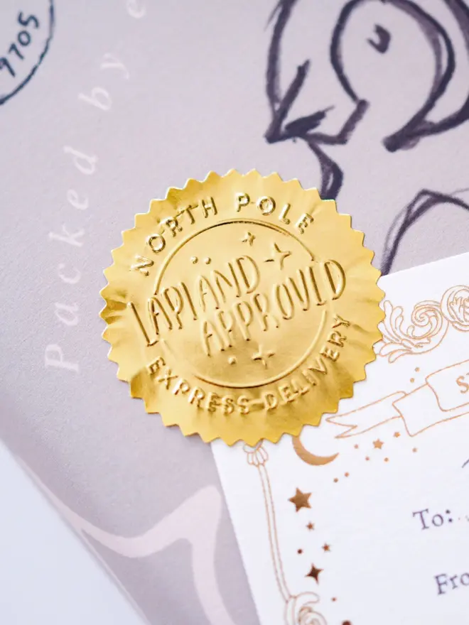 Close-up of an embossed gold seal with scalloped edges and "Lapland Approved" hand-lettering. It seals a lilac parcel next to a gold foil "Special Delivery" label.