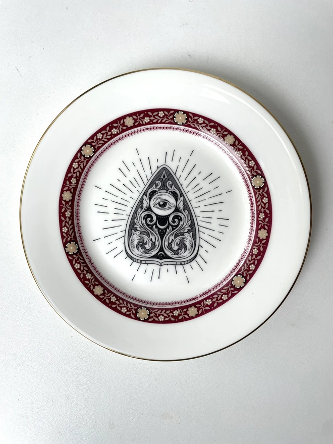 vintage plate with an ornate border, with a printed vintage illustration of a Ouija planchette in the middle 