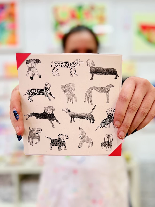 Artist holding a card featuring hand drawn black dogs arranged in a pattern on a white backgroundA charity card featuring hand drawn black dogs arranged in a pattern on a white background