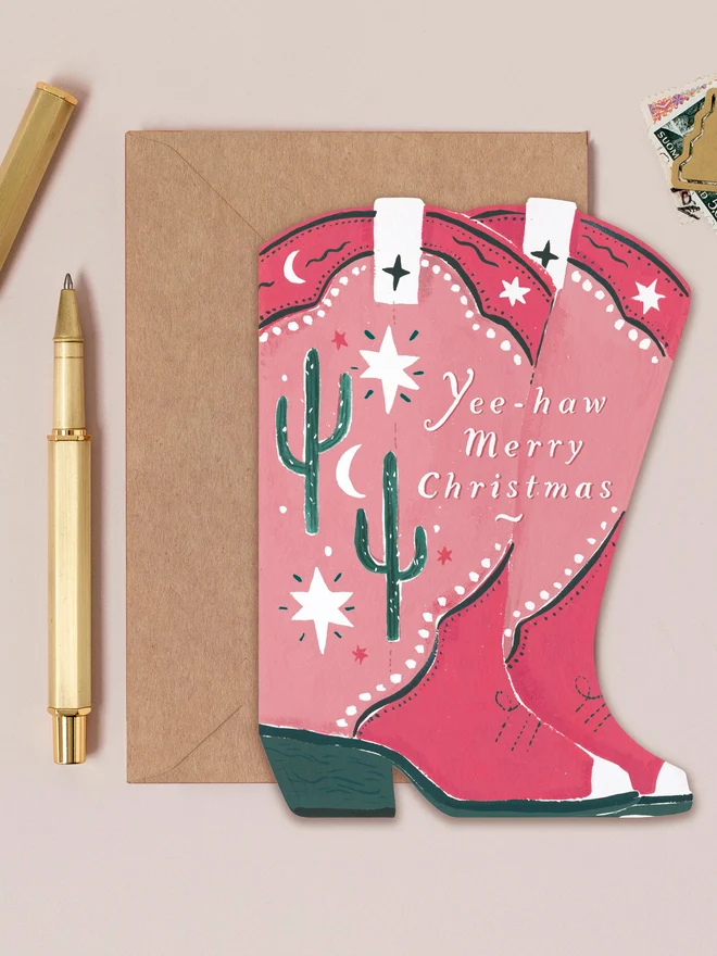 Cowboy Boots Christmas Card by Sister Paper Co, a shaped pink western boots design with a kraft envelope