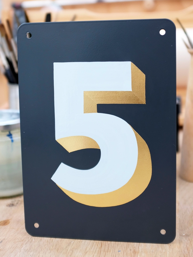 Hand painted pale blue and gold leaf house number 5 on an anthracite grey metal plaque in the artist's studio. 