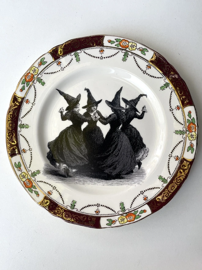 a coven of 4 witches dance hand in hand, the image is victorian in black and white and is printed directly onto an ornate vintage plate