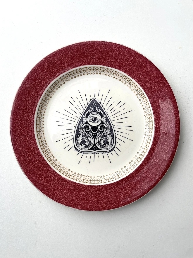 vintage plate with an ornate border, with a printed vintage illustration of a Ouija planchette in the middle 
