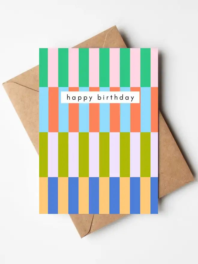 A colourful birthday card featuring a modern abstract striped pattern in green, pink, blue, coral, lilac, yellow and navy, with the words "happy birthday" across the centre. The card is displayed with a kraft envelope.