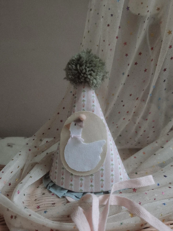 Baby Birthday Hat With Goose, a birthday party hat with a goose. It sits on a wooden hamper. 