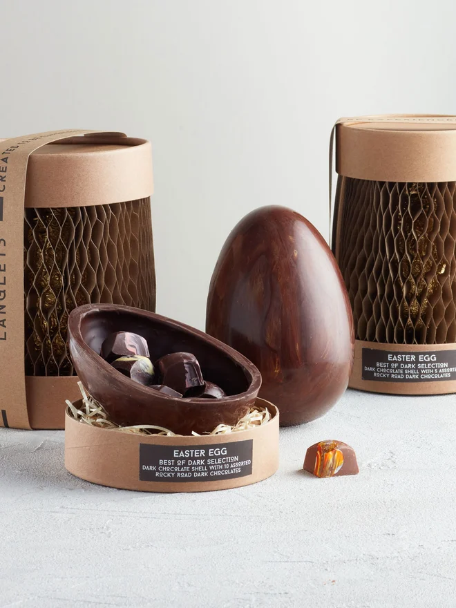 Best Of Dark Chocolate Easter Egg, a chocolate egg sitting on a white surface. It is accompanied by another chocolate egg that is opened showing the chocolates within. 