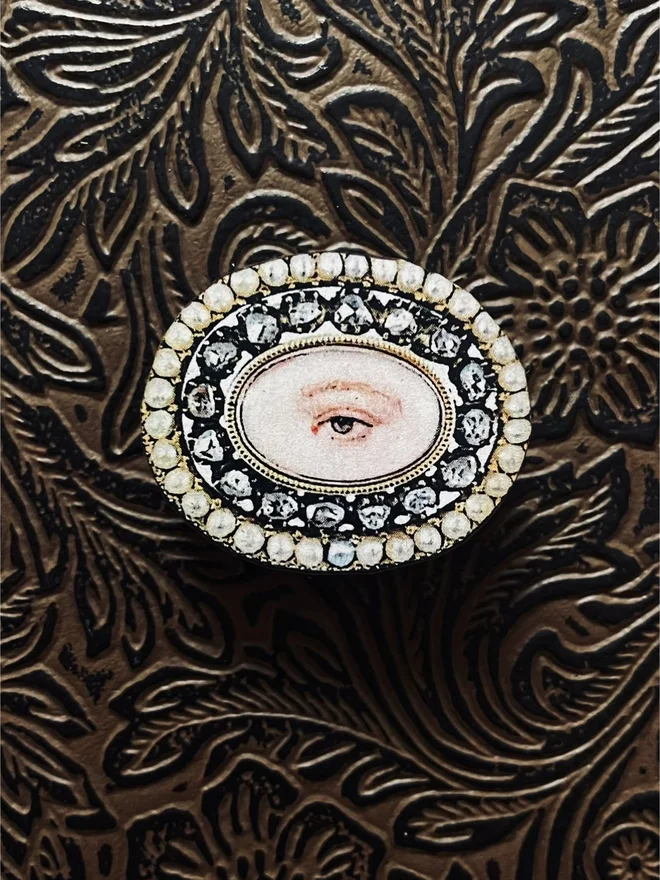 lovers eye brooch, the front is an illustration of a lovers eye with a snake like metal border, printed onto a wood brooch base, with a brass clasp at the back