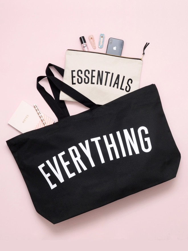 an oversized black canvas tote bag with everything slogan laying on a pink background with an essentials pouch spilling out