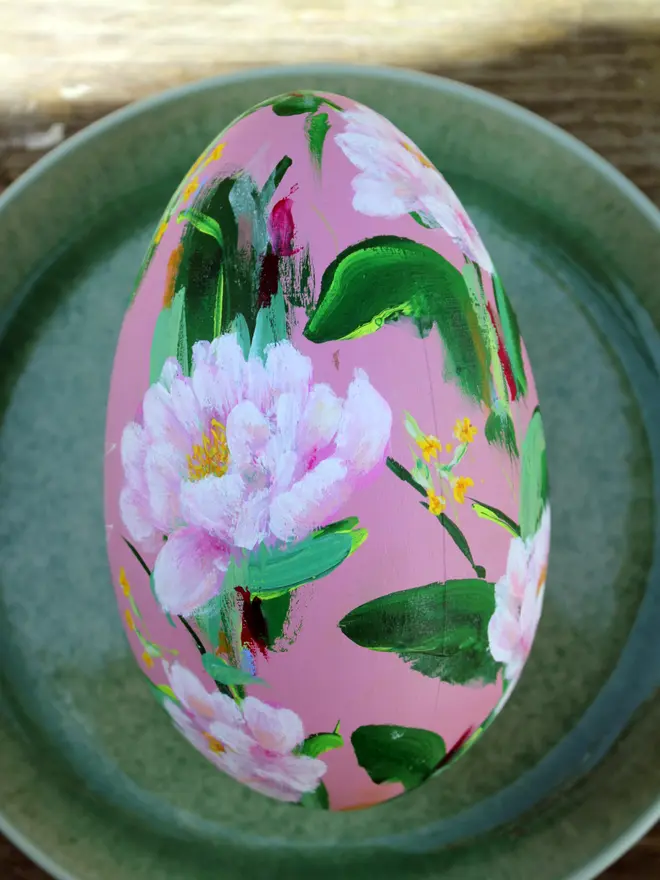 pale pink floral hand painted wooden egg, adorned in pink peonies and yellow cowslip flowers, positioned on a green plate