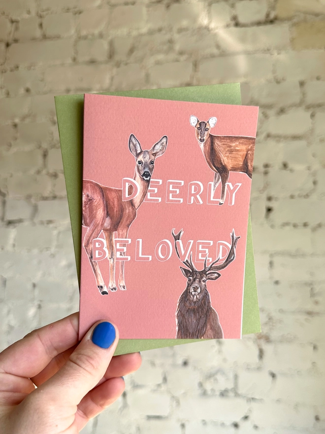 a peachy pink greetings card featuring three illustrations of different deer breeds with the text “deerly beloved” with a green envelope