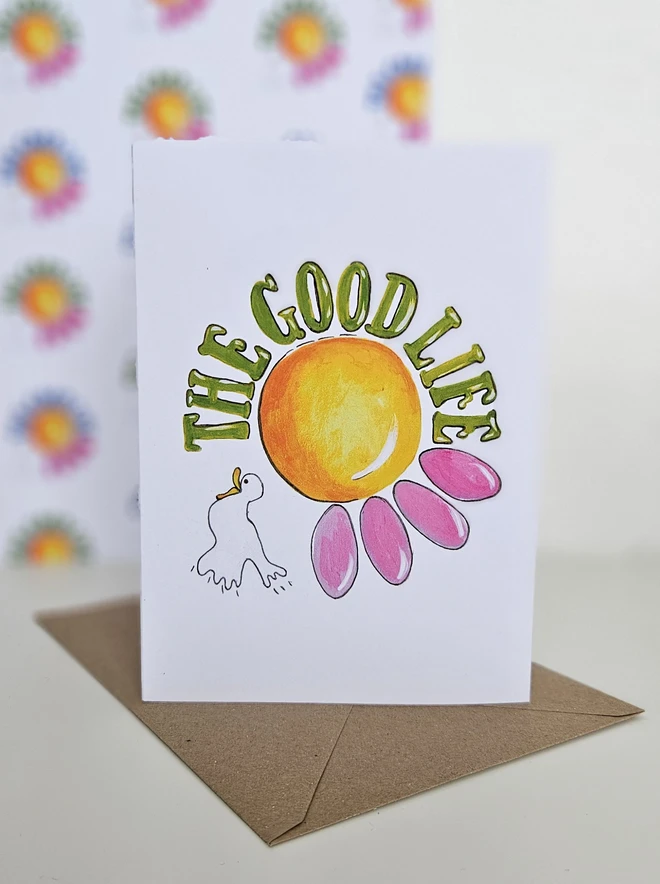 A white card with an illustration of a pink flower and a white bird with The Good Life written using a retro style font in green. 