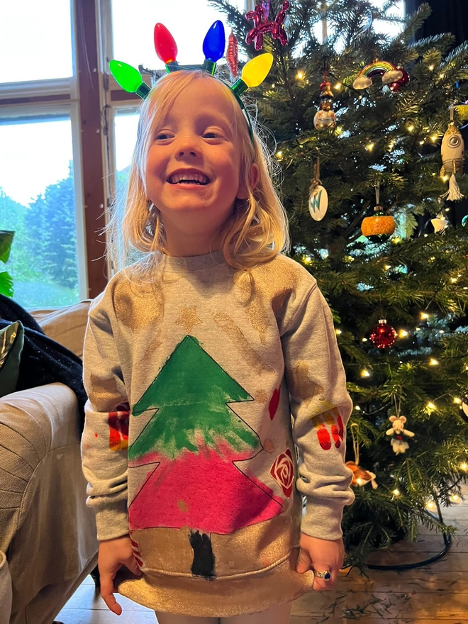 Young girl stood in front of xmas tree wearing her own xmas sweat that she's designed