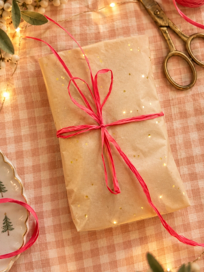 a pretty gift wrapped package laid on a gingham table cloth. the present is wrapped in cream tissue paper with gold flecks and tied with coral rafia there is a christmas gift wrap feel