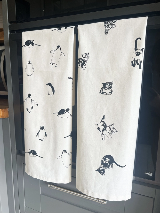 Fold multi cat tea towel next to a multi penguin tea towel hanging on a oven door