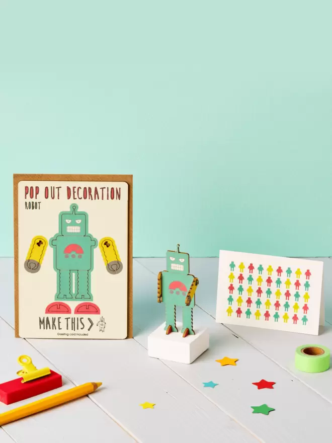 3D green robot decoration and robot pattern greeting card and brown kraft envelope on top of a grey desk in front of a light green coloured background
