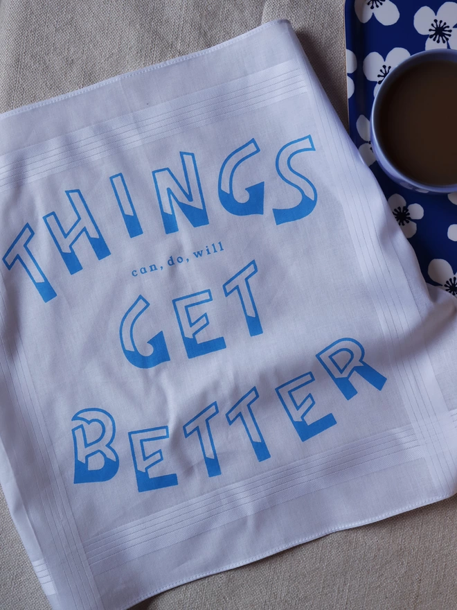Things Get Better handkerchief printed in sky blue on a linen tablecloth alongside a cup of tea on a blue floral tray