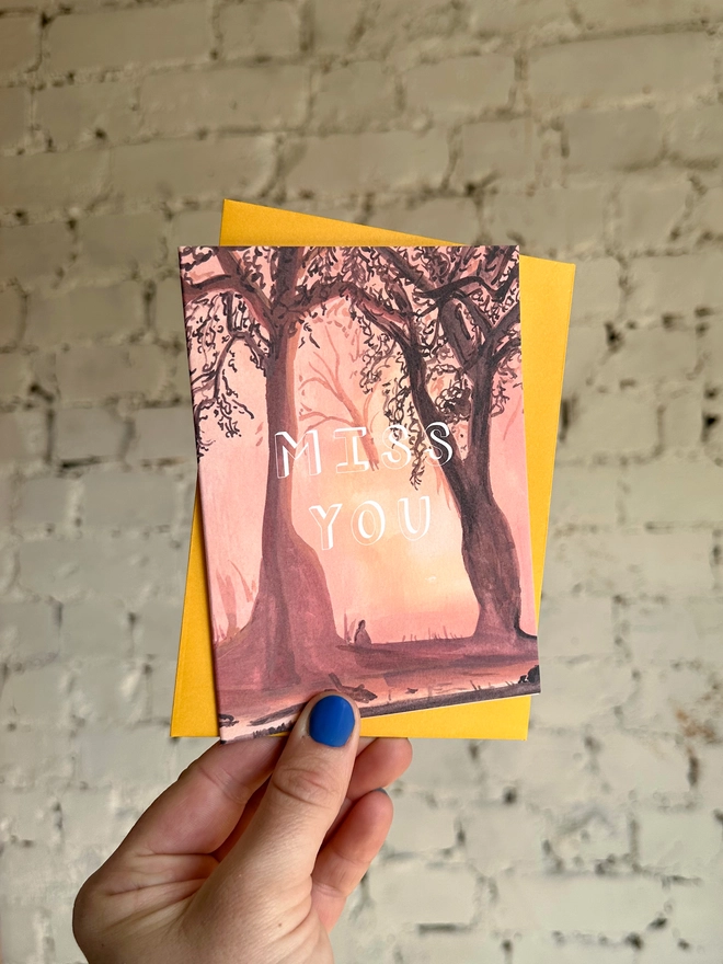 a greetings card featuring an illustration of a sunset through the trees with the text “miss you” with a deep yellow envelope