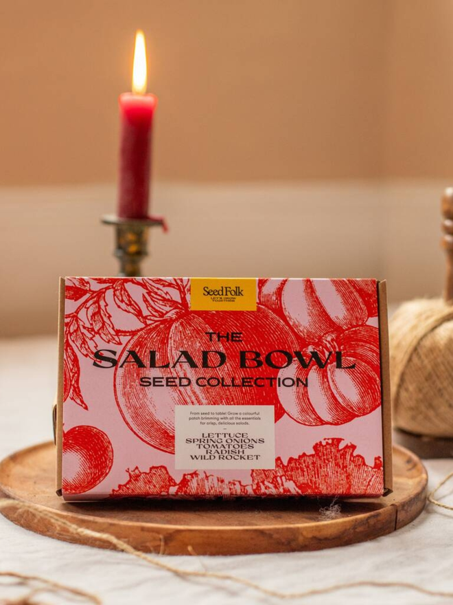the salad bowl seed collection. box in pink and red packaging with illustrated tomatoes photographed in wooden dish with a red candle in the background. 