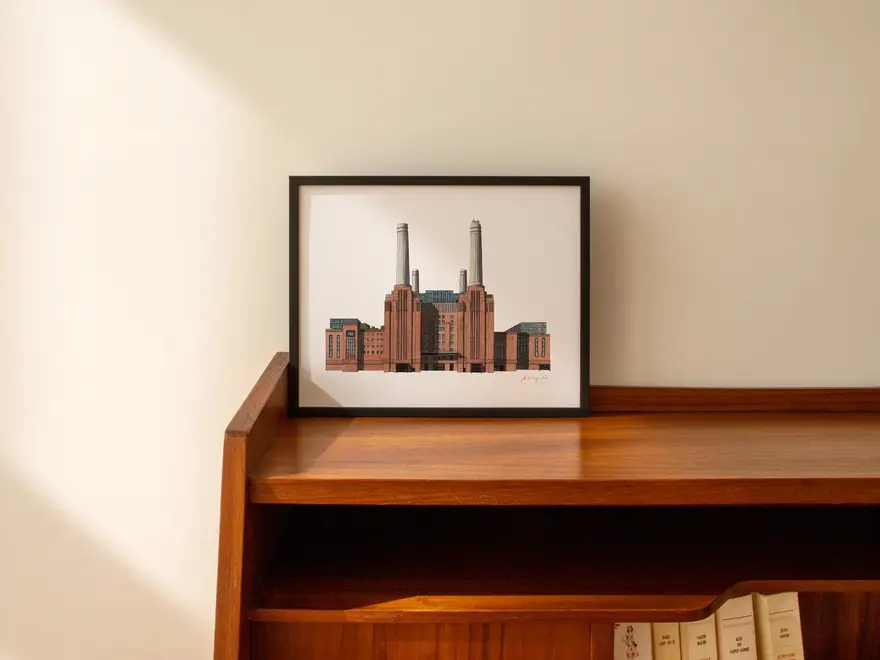Battersea Power Station Giclée Print (Unframed), a print of Battersea Power Station sitting in a black frame against a plain backdrop on a wooden surface. 