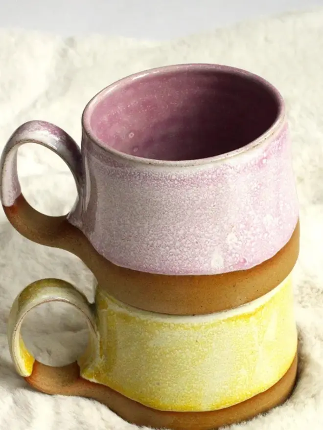 Pink and yellow handmade ceramic cups stacked on top of each other on a soft cream-colored fur background.