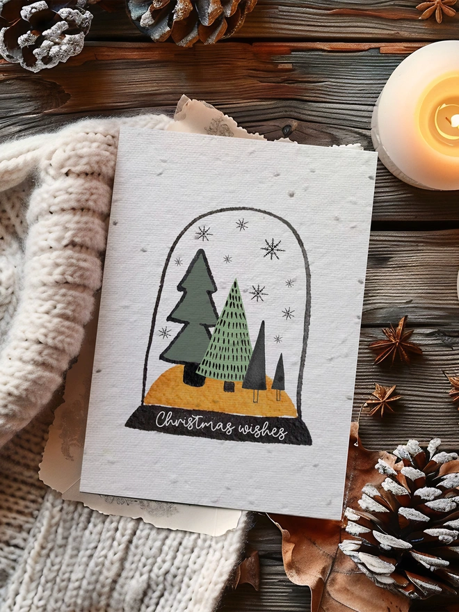Christmas Card with an illustrated snow globe in the centre with trees and 'Christmas Wishes'. The Card is on a dark wood background surrounded by pine cones, a lit candle and a blanket.