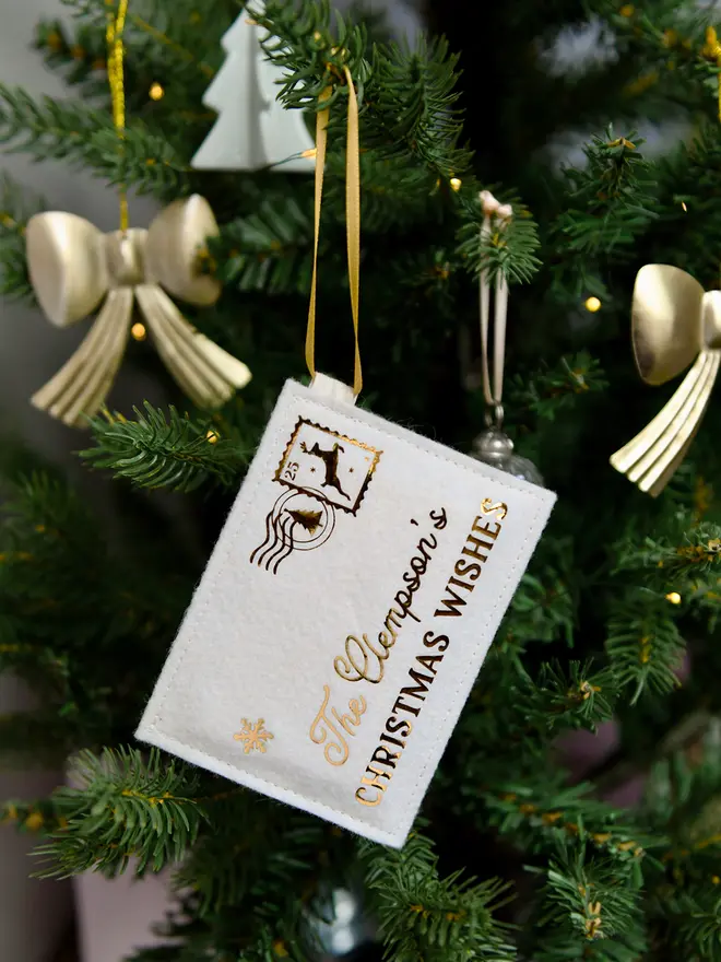 A handmade felt envelope with golden words reading "The Clempson's Christmas Wishes" and a reindeer design stamp in the corner, hangs on a decorated Christmas tree.