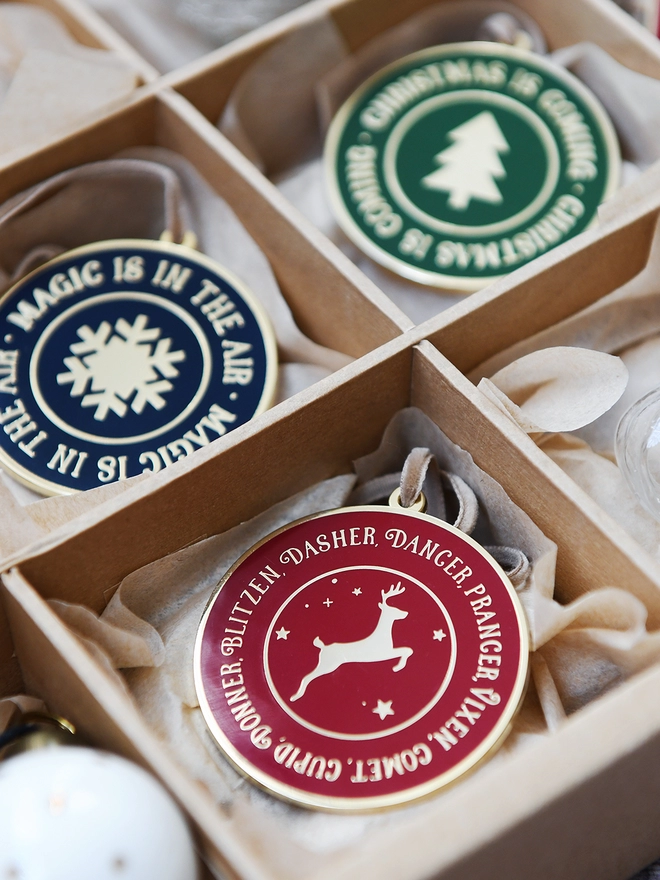 Three enamel Christmas decorations are tucked into a sectioned box. Each decoration has a magical look with festive words on in gold.