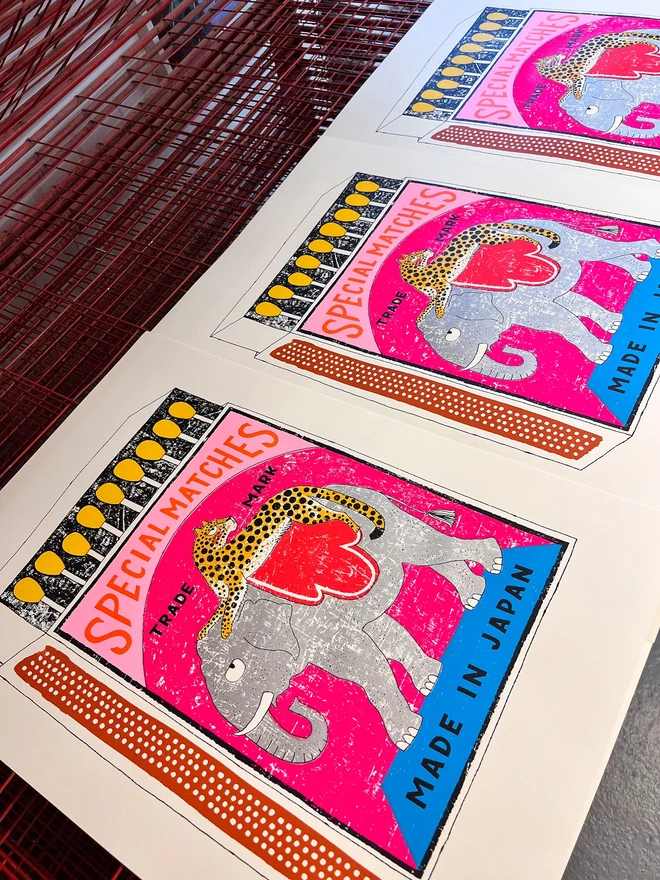 Elephant And Leopard Matchbox Taken For An Extra Long Rid, a colourful matchbox design in the centre of a white card. It sits on a studio rack. 