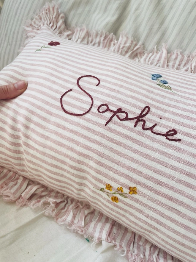     Personalised pink striped stonewashed linen cushion with ruffle trim, hand-painted ditsy florals and hand-embroidered name, styled on a child’s bed.
