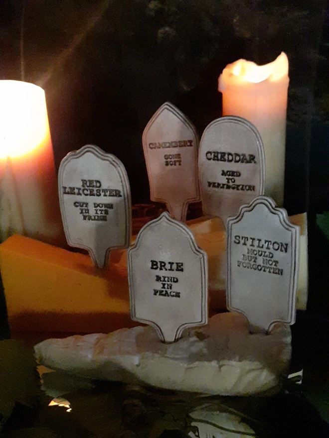 Gothic cheeseboard display featuring five ceramic cheese gravestones among wedges of Cheddar and Brie, warmly lit by two candles.