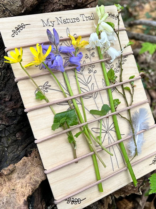 Birch wood nature trail weaving board decorated with artificial wildflowers and leaves, held in place by cream-colored cord, displayed outdoors against mulch and stones.