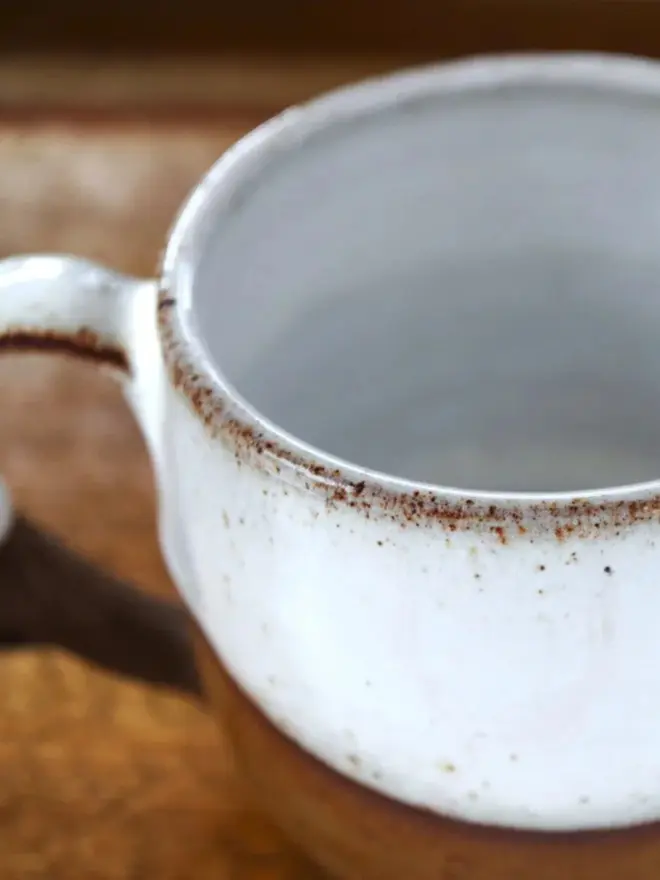 Rustic ceramic mug with a white speckled upper glaze and brown lower half, featuring the word “Brave” on the front, sitting on a wooden table against a neutral background.