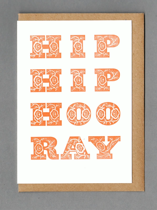 White card with chunky orange text reading 'HIP HIP HOORAY' with a brown envelope behind it