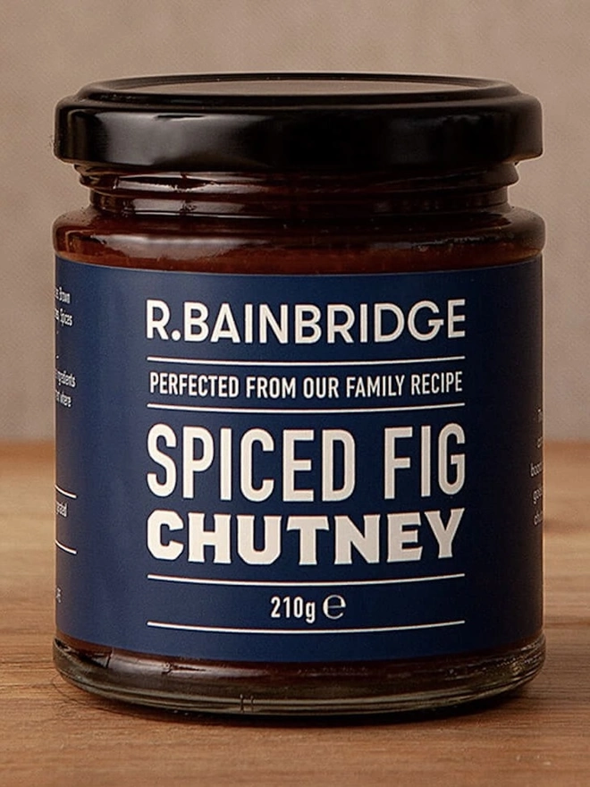 Spiced Fig Chutney, a jar of chutney sitting on a wooden surface against a plain backdrop. 