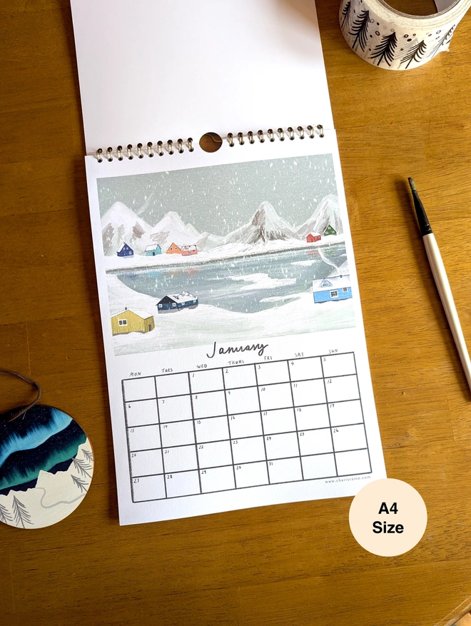 January page of calendar depicts wintry Greenland scene with colourful cabins across a lake.