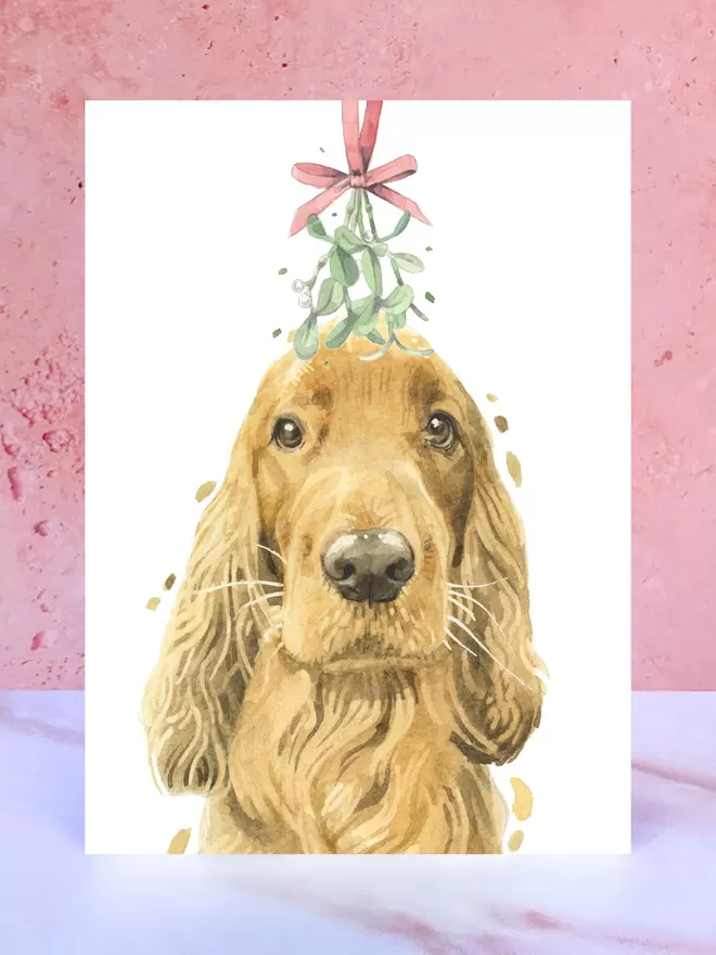 A Christmas card featuring a hand painted design of a Ginger Cocker Spaniel, stood upright on a marble surface.
