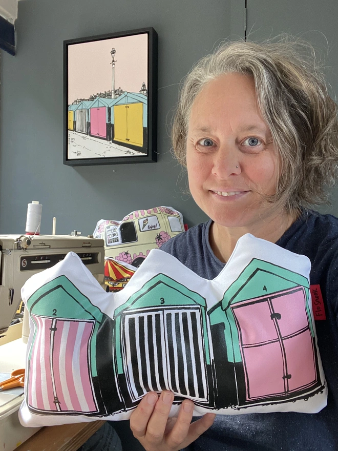 Row of beach huts cushion handmade by the artist Flo Snook in her studio. A sea-green and pink cushion held in the hand of Flo Snook next to her sewing machine with an original beach huts painting on the wall behind