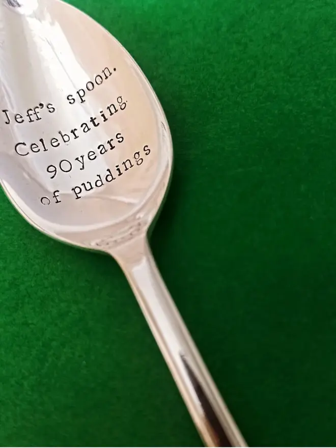 A vintage silver related dessert spoon. Handstamoed with Geoff's spoon Celebrating 90 years of puddings. The spoons is on a green background  
