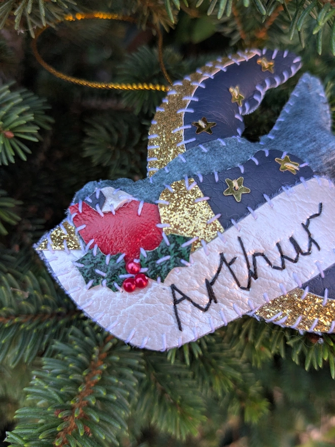Hand stitched personalised swallow Christmas decoration made from vegan-friendly faux leather and velvet, featuring gold stars, festive holly and a cream banner personalised with a name, hanging from a gold cord on a Christmas tree.